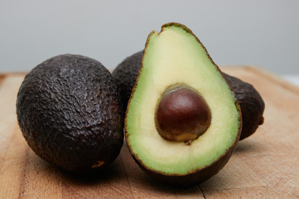 A trio of avocados is displayed on a rustic wooden board, their deep green skins contrasting with the warm tones of the wood. One avocado has been sliced open, revealing its vibrant, creamy interior and a large brown seed in the center.