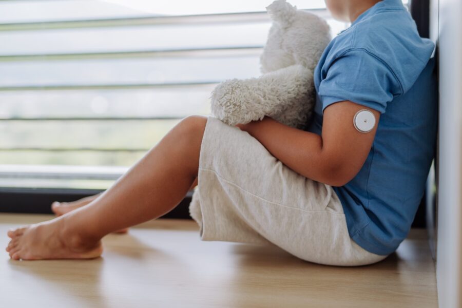 Diabetic boy with a continuous glucose monitor sitting by the window, holding his stuffed teddy bear and looking outside.