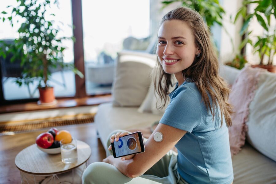A young girl sits on a couch, holding a Continuous Glucose Monitoring (CGM) device against her arm. Sunlight casts a warm glow on lush green plants in the background. Spring Cleaning Your Diabetes Routine Featured Image