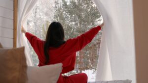 Woman looking out of a snowy window while sitting on a bed