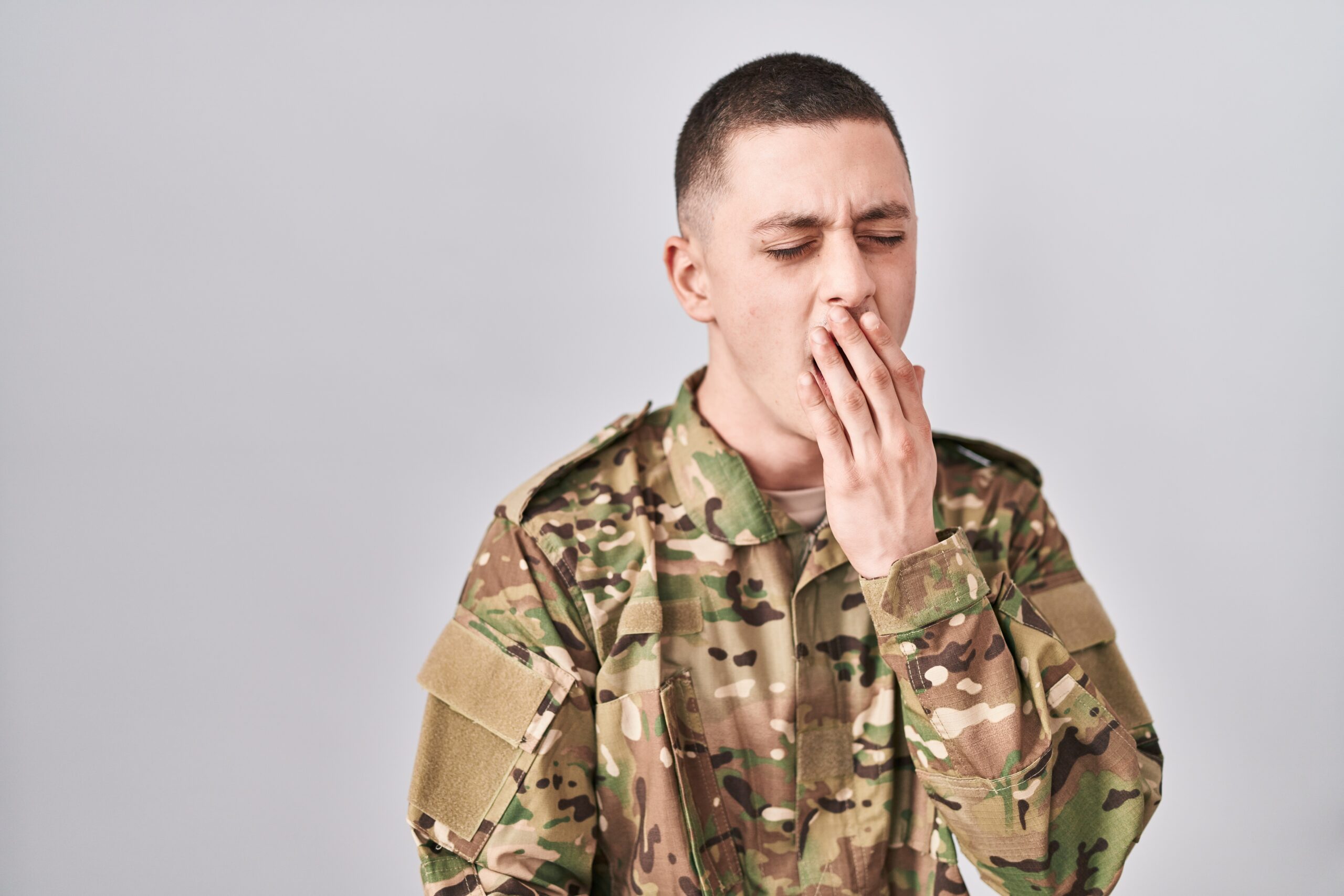 Young man wearing camouflage army uniform bored yawning tired covering mouth with hand. restless and sleepiness.
